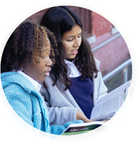 Two adolescent girls standing together as they review a textbook.